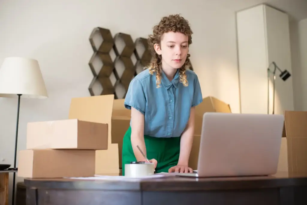 En ung kvinna med uppsatt lockigt hår, iklädd en blå skjorta med knappar, står vid sitt matbord med staplade kartonger och kontrollerar sin laptop för att hantera dropshipping-beställningar hemifrån.
