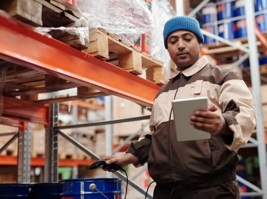 En manlig lagerarbetare i blå mössa och brunbrun arbetsuniform håller en surfplatta för att skanna och kontrollera lagret på höga orangea industriella hyllor fyllda med kartonger och förpackade varor i ett BLB Dropshipping-distributionscenter.
