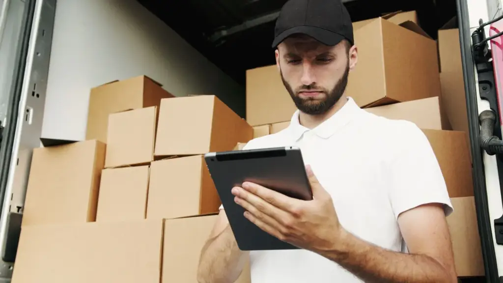 En skäggig man i vit pikétröja och svart keps, håller i en surfplatta för att kontrollera orderuppgifter, står framför staplade kartonger redo för leverans i ett dropshipping-distributionslager.